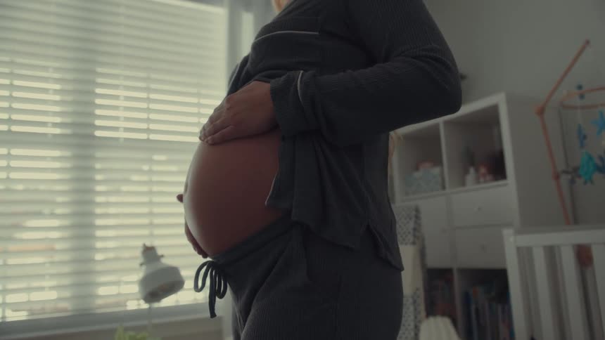 A close view of a pregnant woman wearing grey pajamas as she stands in a nursery and gently places both hands on her bare stomach while looking down in profile