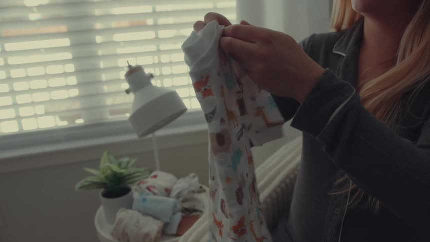 A pregnant woman sitting indoors folding baby clothes and placing them on a table beside her