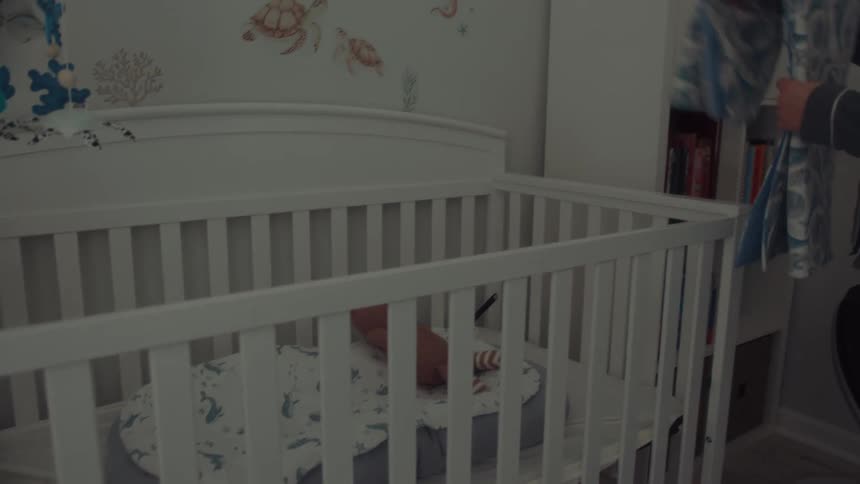A pregnant woman wearing grey pajamas stands in a nursery as she places a stuffed animal into a white crib and drapes a blue blanket over the side
