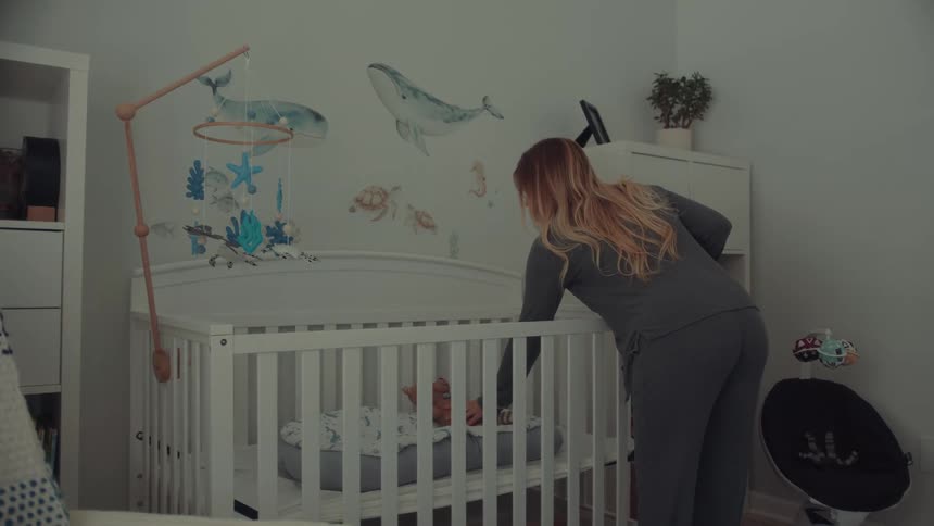 A woman wearing gray pajamas enters a nursery and places a blanket inside a white wooden crib before folding another blue blanket near a white bookshelf and whale wall decals