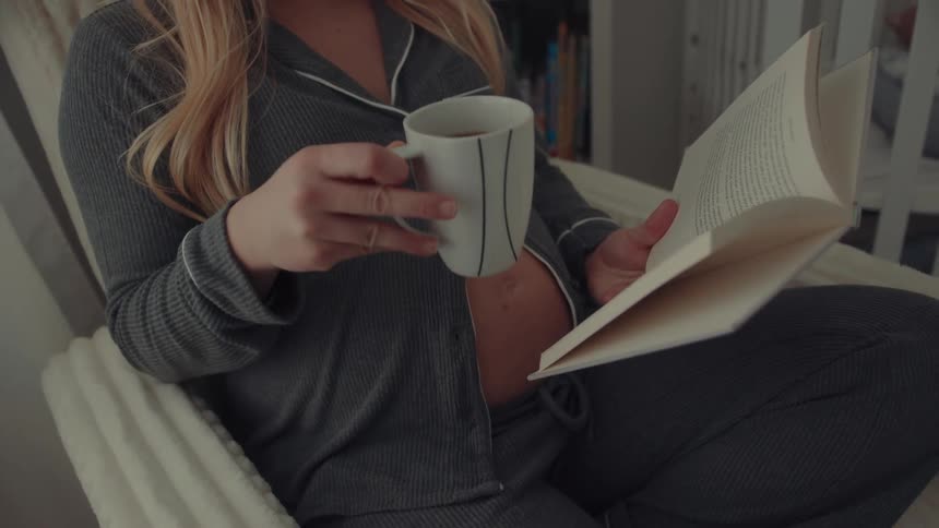 A close view of a pregnant woman with long blonde hair wearing charcoal ribbed pajamas while sitting and reading a book with her bare stomach partially visible