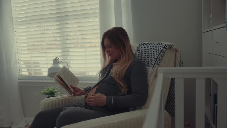 A pregnant woman wearing grey pajamas sits in a cream colored rocking chair in a nursery while reading a book and resting her hand on her stomach