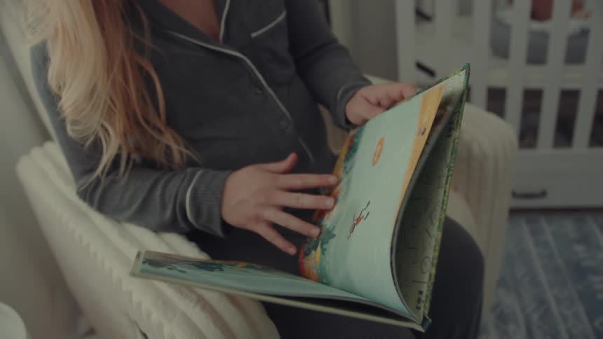 A pregnant woman wearing pajamas sitting in an armchair in her bedroom reading a storybook