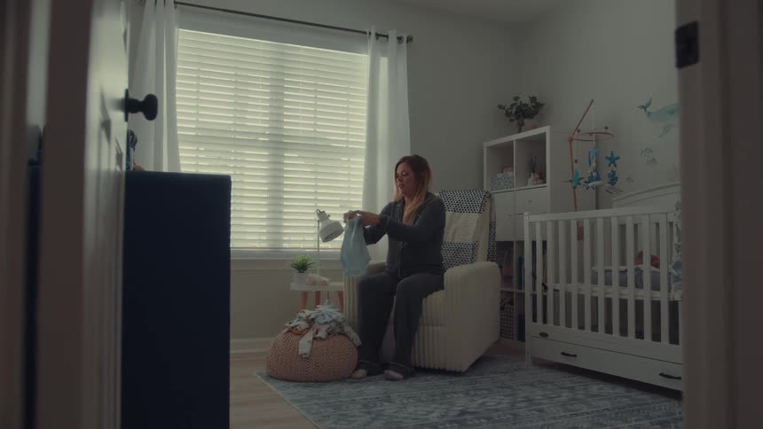 A pregnant woman wearing grey pajamas sits in a cream colored rocking chair inside a nursery as she folds small blue garments while a white crib stands nearby