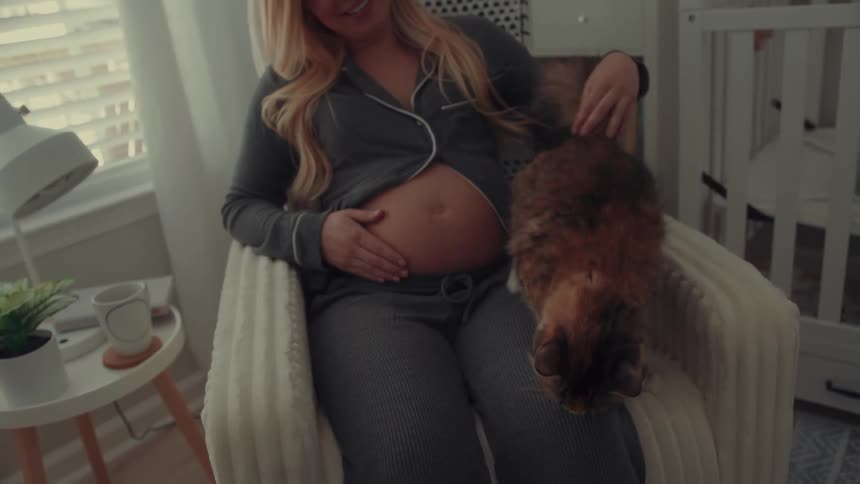 A pregnant woman wearing grey pajamas sits in a white rocking chair and smiles while petting a calico cat in a nursery with a crib and window in the background