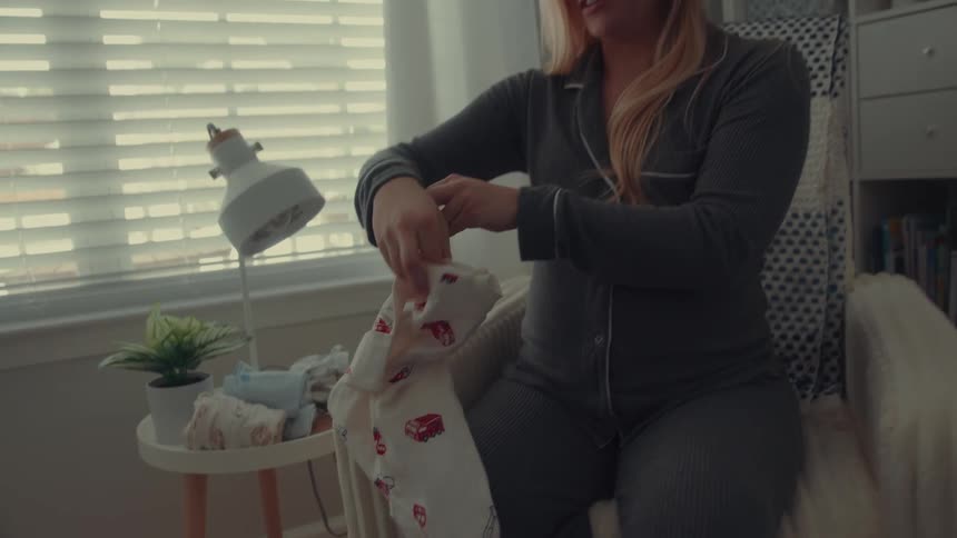 A pregnant woman sits in a chair and examines a white baby onesie with a red pattern she carefully folds the garment and places it onto a small side table next to other baby clothes