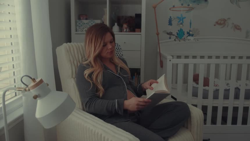 A pregnant woman sitting in a chair drinking a cup of coffee and reading a book gently rubbing her baby bump with a crib beside her