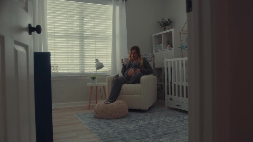 A pregnant woman sits in a rocking chair in a nursery drinking from a mug and looking out the window the room features a white crib a blue patterned rug and a small side table with a lamp