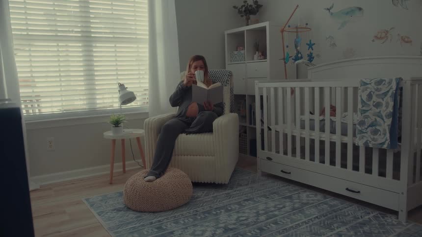 Pregnant woman reads a book sitting in an armchair next to a crib