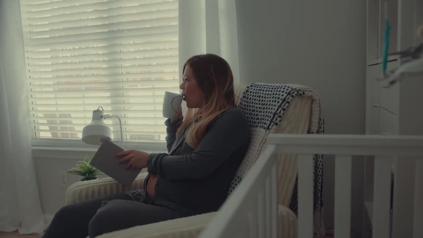 A pregnant woman sits in a rocking chair in a nursery reading a book while resting her hand on her stomachshe pauses her reading to take a sip from a mug before continuing to look through the pages
