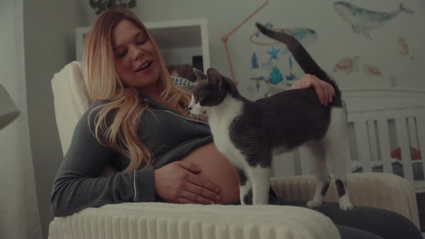 A pregnant woman wearing gray pajamas sits in a rocking chair and smiles while petting a gray and white cat that stands on her lap in a nursery with a white crib