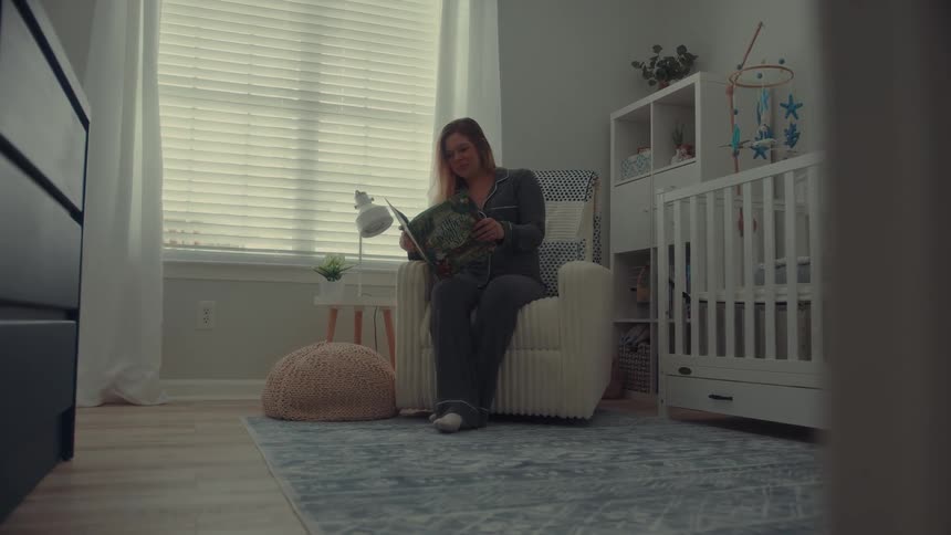 A woman wearing grey pajamas sits in a cream colored rocking chair in a nursery and reads a green book while holding a mug near a white crib