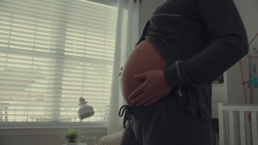 A pregnant woman stands in a nursery gently cradling her bare belly with both hands while smiling the room is filled with soft natural light coming through a window with blinds as she looks down at her stomach