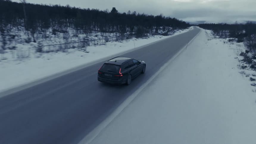Black car driving on a road surrounded by snow and trees under a cloudy sky