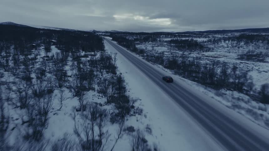 An aerial view shows a car driving on a road through a snowy landscape with many small trees under a dark cloudy sky
