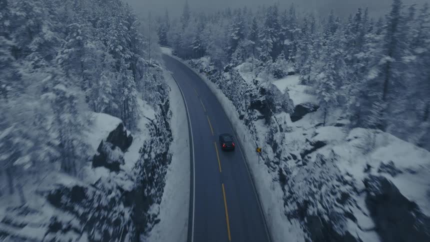 Car drives along winding road in snowy forest seen from above