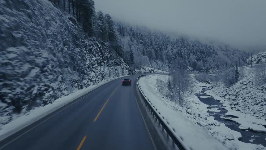Car drives along winding road in snowy mountain landscape with river beside