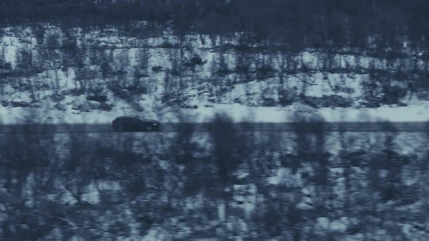 A dark suv travels along a lonely snowdusted highway that cuts through a dense thicket of bare winter treesthe aerial perspective captures the vehicle moving steadily across a cold monochromatic landscape of hills and frozen brush