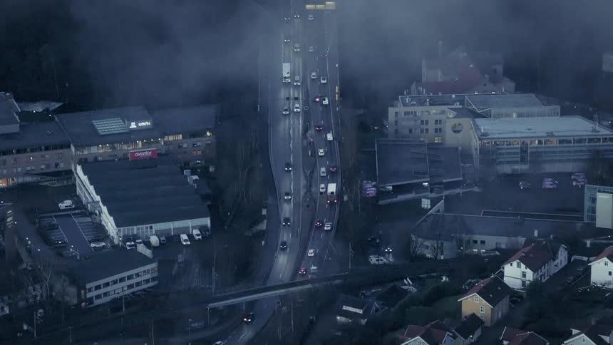 An aerial view shows cars and trucks driving along a highway through a city where a low layer of fog hangs over the buildings and trees during the evening
