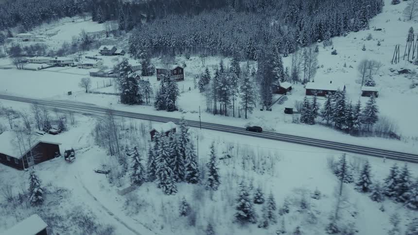 An aerial view shows a dark car driving along a road through a snowy valley with scattered houses and a dense forest of evergreen trees under an overcast sky