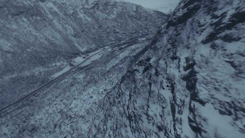 An fpv drone captures a highspeed descent down a rugged snowcovered mountain face toward a valley floor the flight levels out over a desolate winter landscape featuring a winding road and sparse leafless trees under a grey sky
