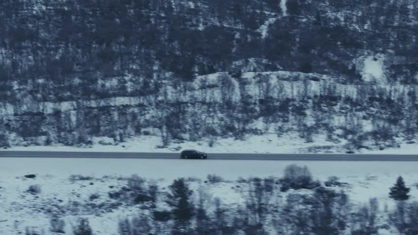 An aerial camera tracks a dark vehicle as it drives across a vast snowcovered landscapethe road stretches horizontally across the frame positioned between a dense hillside of bare trees and a foreground of frozen brush