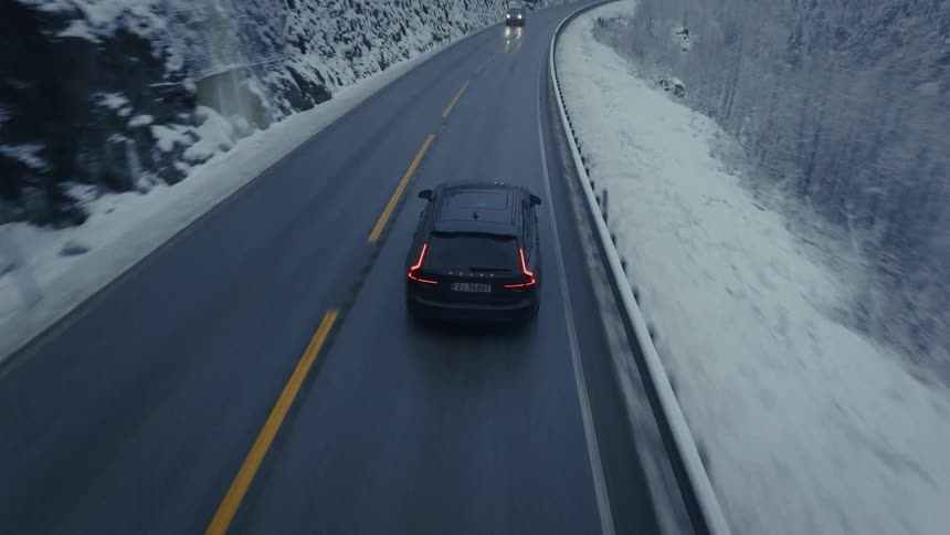 An aerial drone shot follows a dark suv as it travels along a winding mountain road flanked by snowcovered cliffs and trees the overcast sky and damp pavement create a moody wintry atmosphere throughout the drive