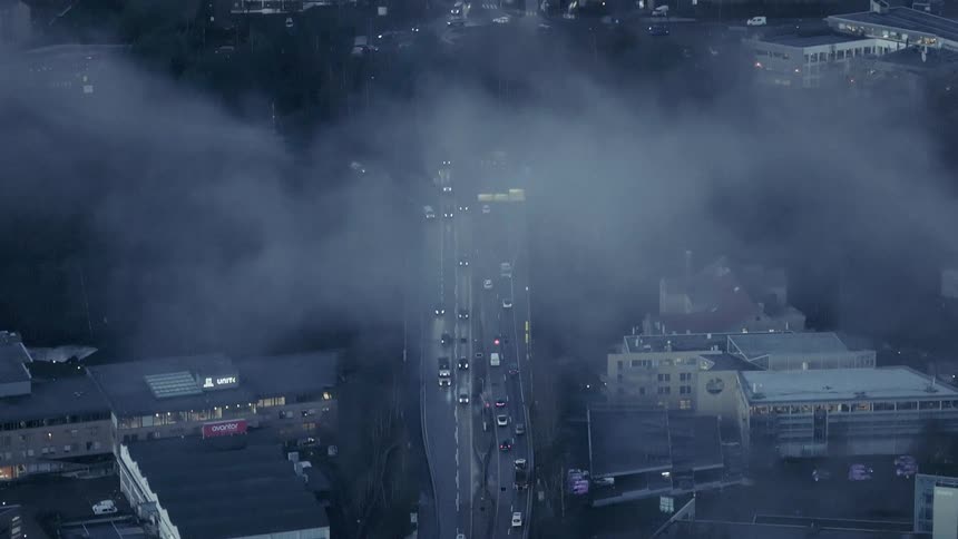 An elevated view shows many cars driving on a busy city highway and through a traffic circle as thick white fog rolls over the surrounding buildings and streets during the evening