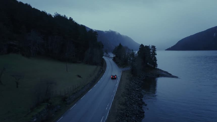 Car driving along a narrow road between a lake and mountains under a cloudy sky