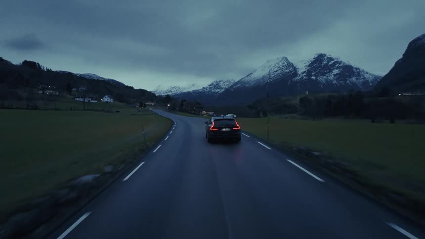 The video features a dark car driving along a winding twolane road through a mountainous landscape at duskas the car moves toward snowcapped peaks in the distance its red taillights remain visible against the bluetoned lowlight environment