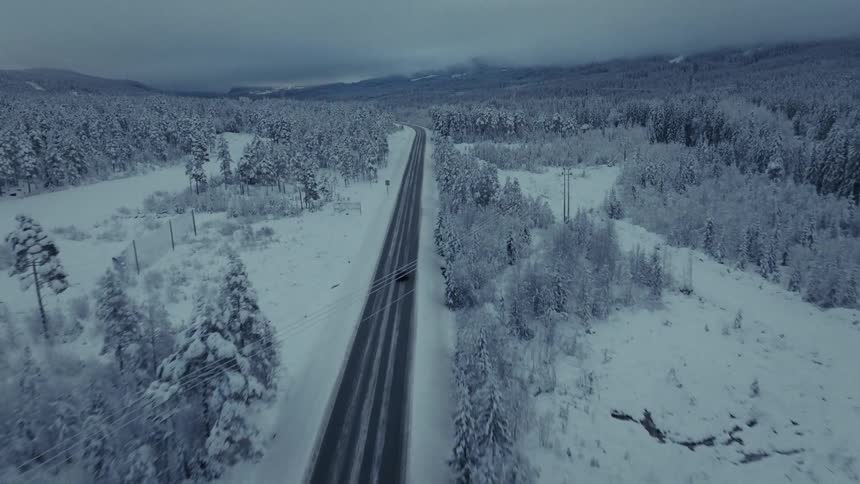 An aerial view shows a car as it drives on a straight road through a vast forest covered in deep snow under a dark cloudy sky