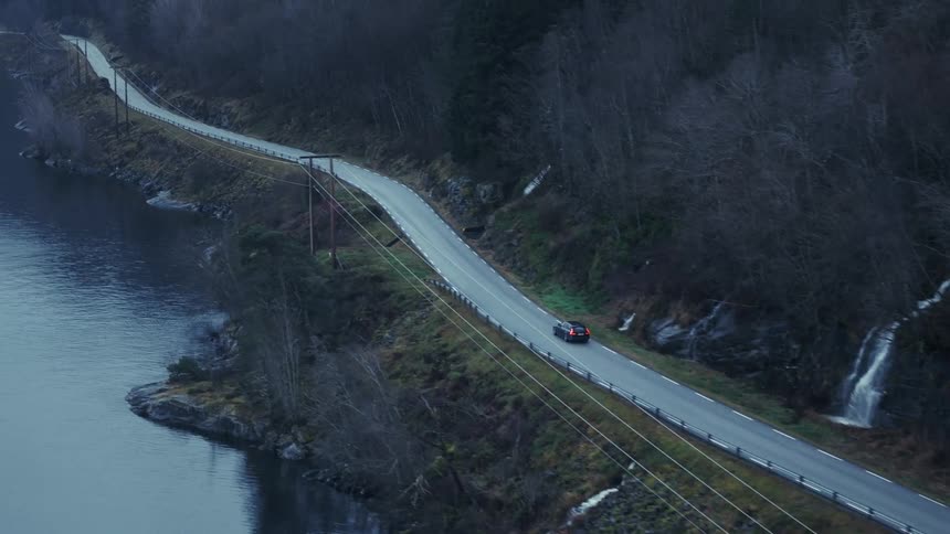 An aerial view shows a dark car as it drives on a winding road that follows the coastline of a lake near a small waterfall on a steep wooded hill under a gray sky