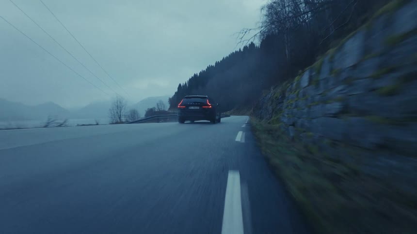 A dark car drives along a winding coastal road at dusk between a steep rocky hillside and a calm body of water under a dim overcast sky