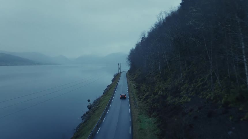An aerial view shows a dark car driving along a narrow road that curves between a steep wooded hillside and a large body of water under a cloudy sky