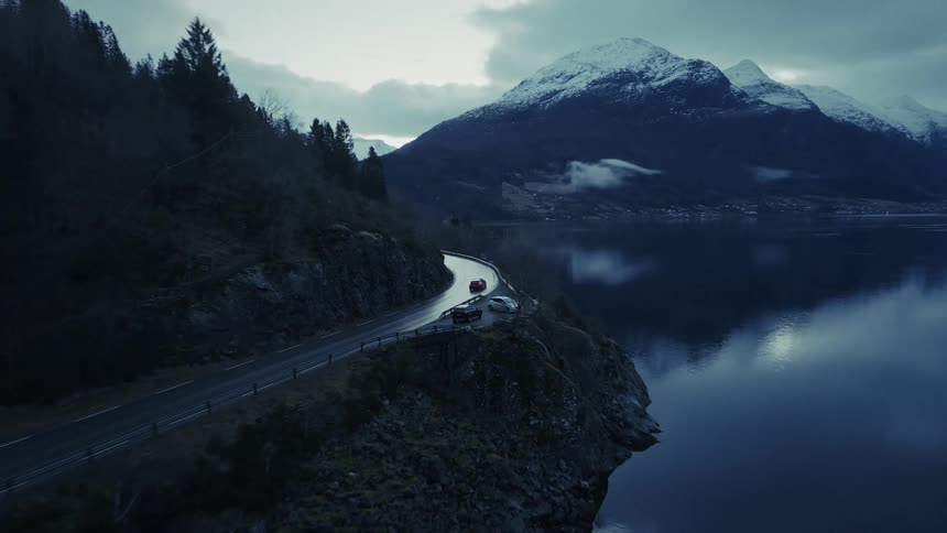 An aerial camera follows a red car as it winds along a scenic coastal road nestled between steep forested cliffs and a calm body of water the moody overcast atmosphere highlights the dramatic snowcapped mountains reflecting in the lake during the twil