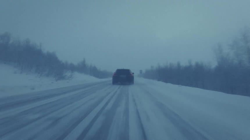 A black car drives down a lonely snow covered highway during a heavy winter blizzard while passing dark trees and blue road signs under a dim overcast sky