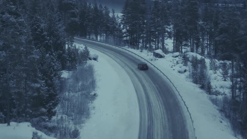 An aerial view captures a dark car driving along a winding snowcovered road through a dense evergreen forest the surrounding landscape is blanketed in thick snow under a dim overcast sky