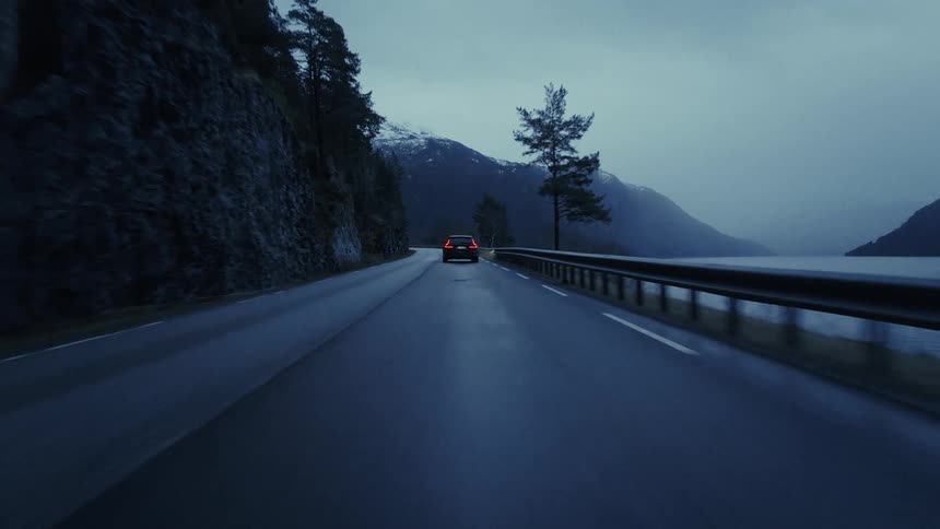 The video shows a black car driving along a winding coastal road at dusk to the left of the road is a steep rocky cliffside covered in dark evergreen trees