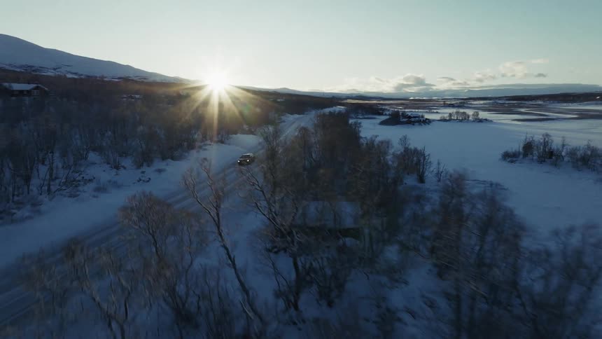The video opens with an aerial shot of a dark car driving down a snowcovered road toward a low bright sun as the camera pans right the car disappears behind a dense thicket of frosted trees shifting the focus to the vast icy landscape