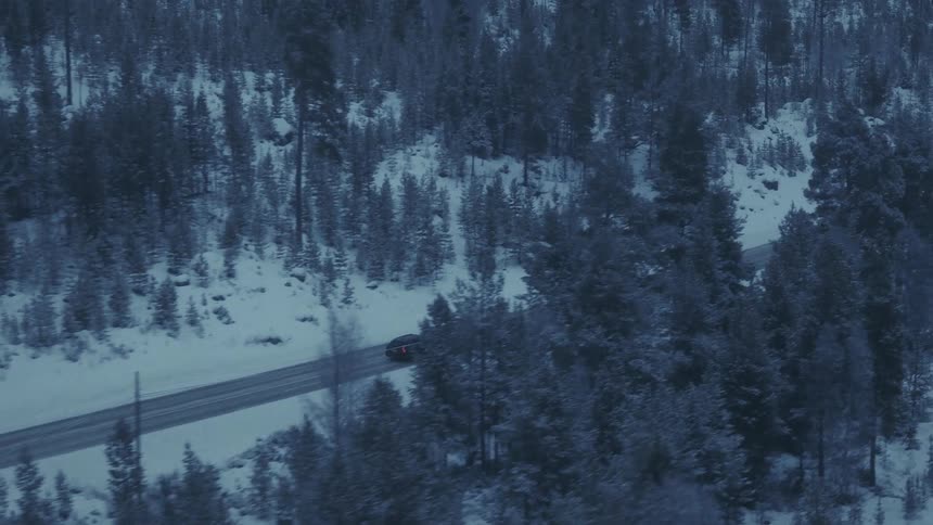 A dark car drives along a remote road winding through a dense forest covered in snow during the blue hour with tall pine trees surrounding the area and mountains visible in the distance