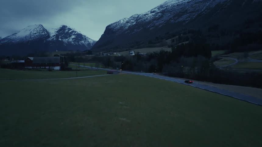 Winding road cuts through green fields toward snowy mountains at dusk
