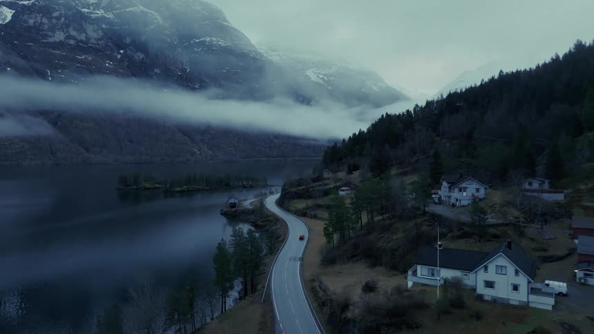 Red car drives along winding road between snowy mountains and lake
