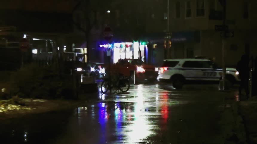 In a nighttime urban setting an nypd vehicle with flashing blue and red lights is parked on a wet reflective streetthe scene is illuminated by bright streetlights showing a few people walking nearby and a red truck driving through the background
