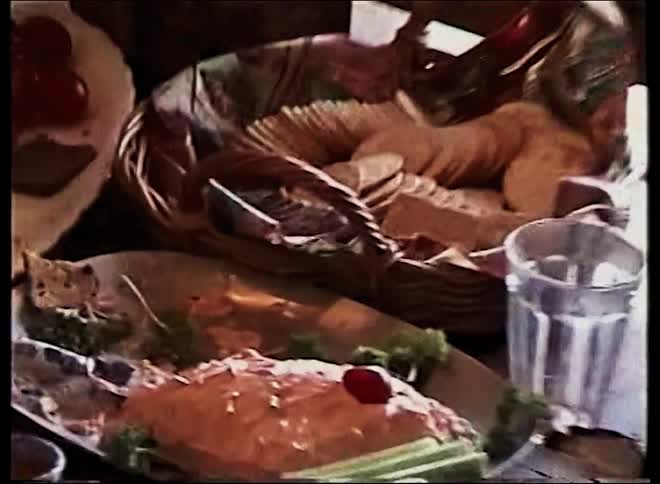 A wooden table is filled with a variety of appetizers including cheese blocks crackers in a basket smoked salmon and various dips served on different plates during a social gathering