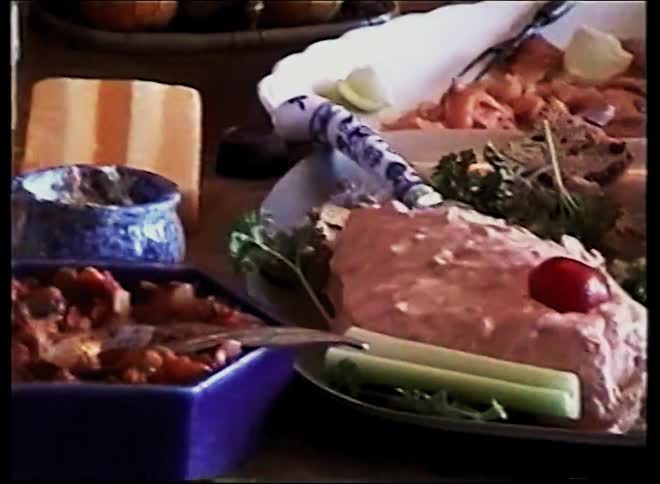 A person uses a slicer to cut a block of cheese on a wooden table near a bowl of dip and a blue dish of relish while other breakfast items like sliced salmon and a bread basket sit arranged on a large platter in the background