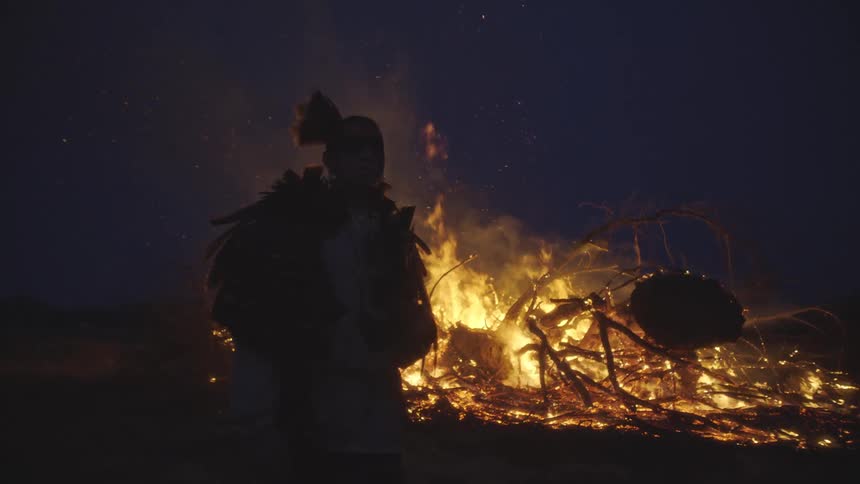 A man in traditional tribal clothing stands stoically in front of a massive bonfire at night as bright orange flames consume dry branches and send sparks into the dark sky