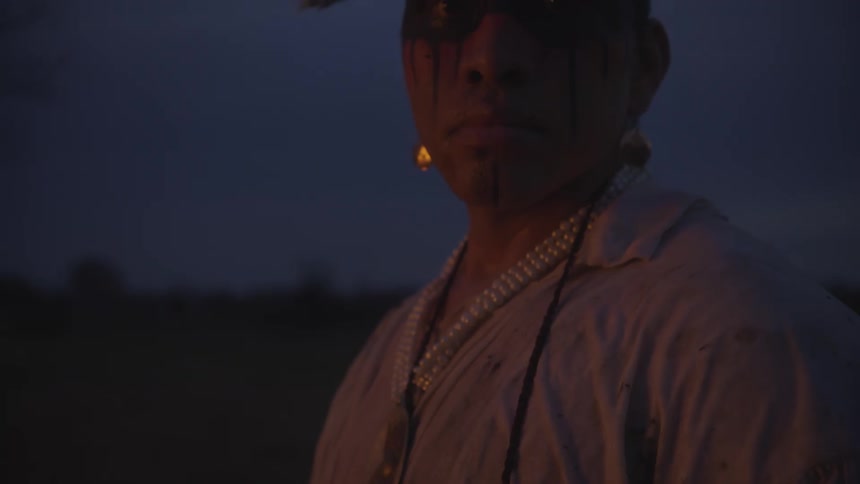 A man wearing traditional feathers and face paint with a gold chest plate and pearl necklaces looks into the camera during a dark dusk or evening setting in an outdoor field