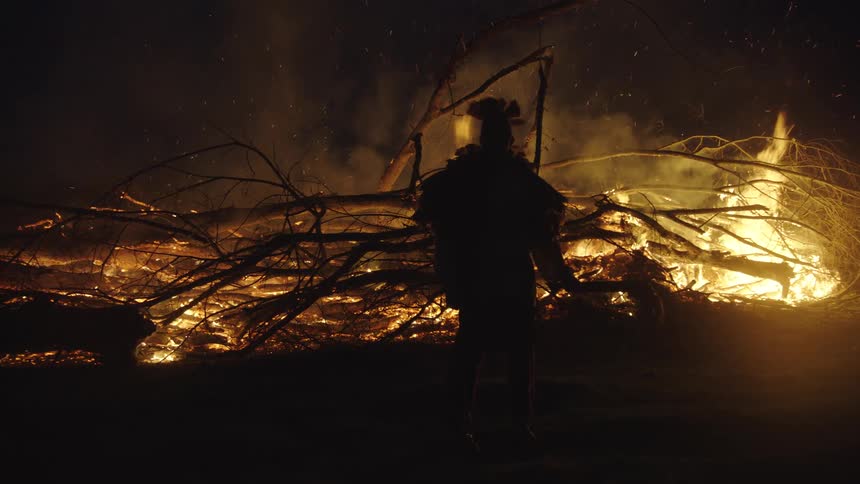 A person in a feathered headdress stands in silhouette before a large bonfire at night as bright orange flames consume branches and send sparks into the dark air
