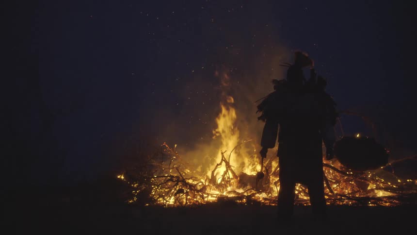 Silhouette of a person standing in front of a bonfire with burning branches at night