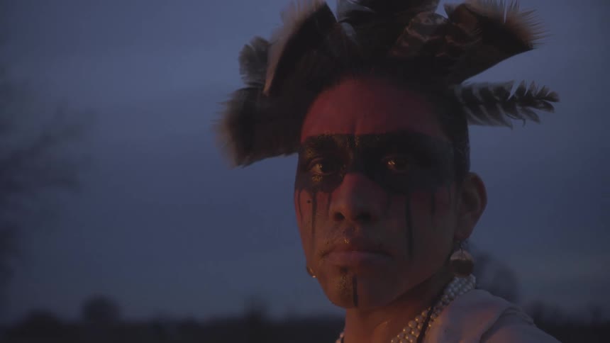 An indigenous man with black face paint and a feathered headdress turns his head to look directly at the camera against a dark twilight sky in an outdoor setting